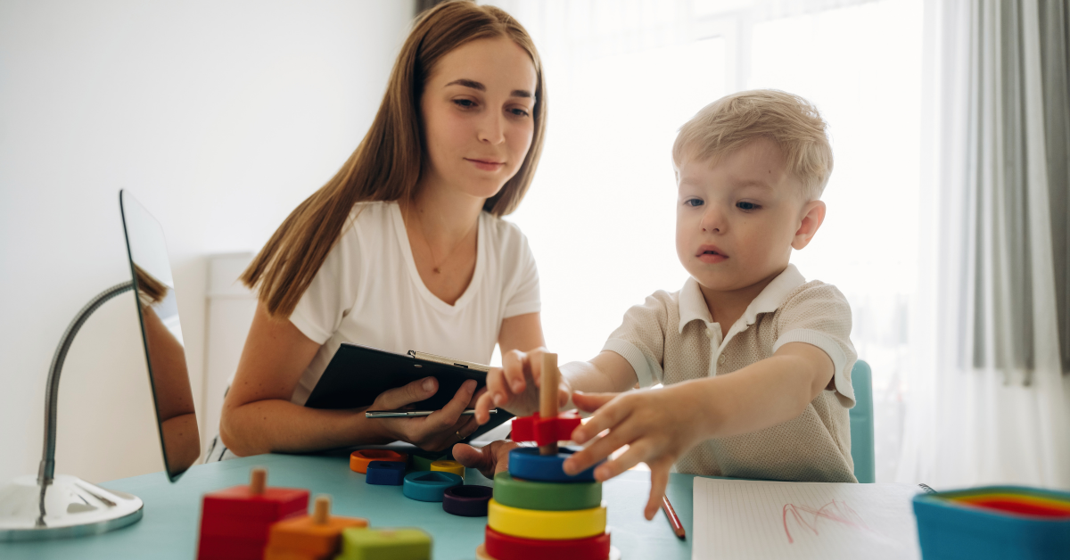banner-autism-services-finding-the-right-fit-how-to-choose-an-ABA-provider-for-your-child (1) young toddler working with a woman ABA provider with toys on a table while she holds a clipboard.