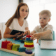 young toddler working with a woman ABA provider with toys on a table while she holds a clipboard.
