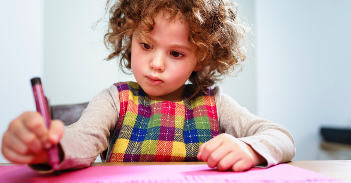 banner-autism-services-supporting-autistic-youth-through-summer-transitions preschool aged child with brown curly hair holding a burgundy crayon concentrating on coloring on a pink paper