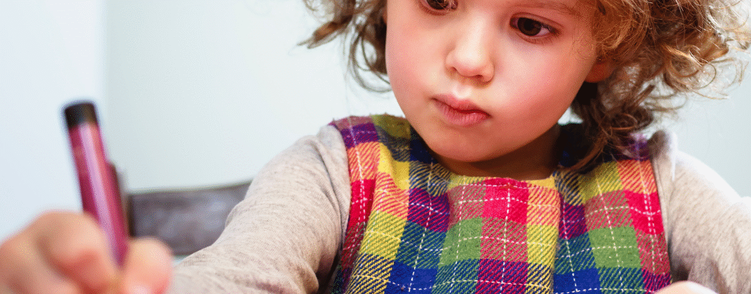 preschool aged child with brown curly hair holding a burgundy crayon concentrating on coloring on a pink paper