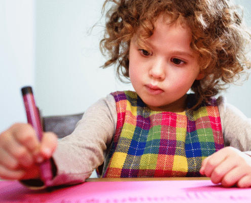 preschool aged child with brown curly hair holding a burgundy crayon concentrating on coloring on a pink paper