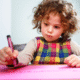 preschool aged child with brown curly hair holding a burgundy crayon concentrating on coloring on a pink paper